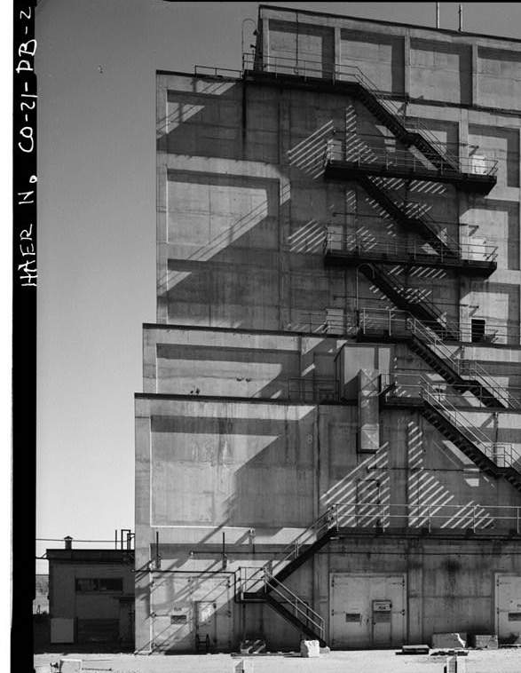 Black-and-white archival photograph of a multi-story concrete industrial building with exterior metal fire stairs and strong diagonal shadows